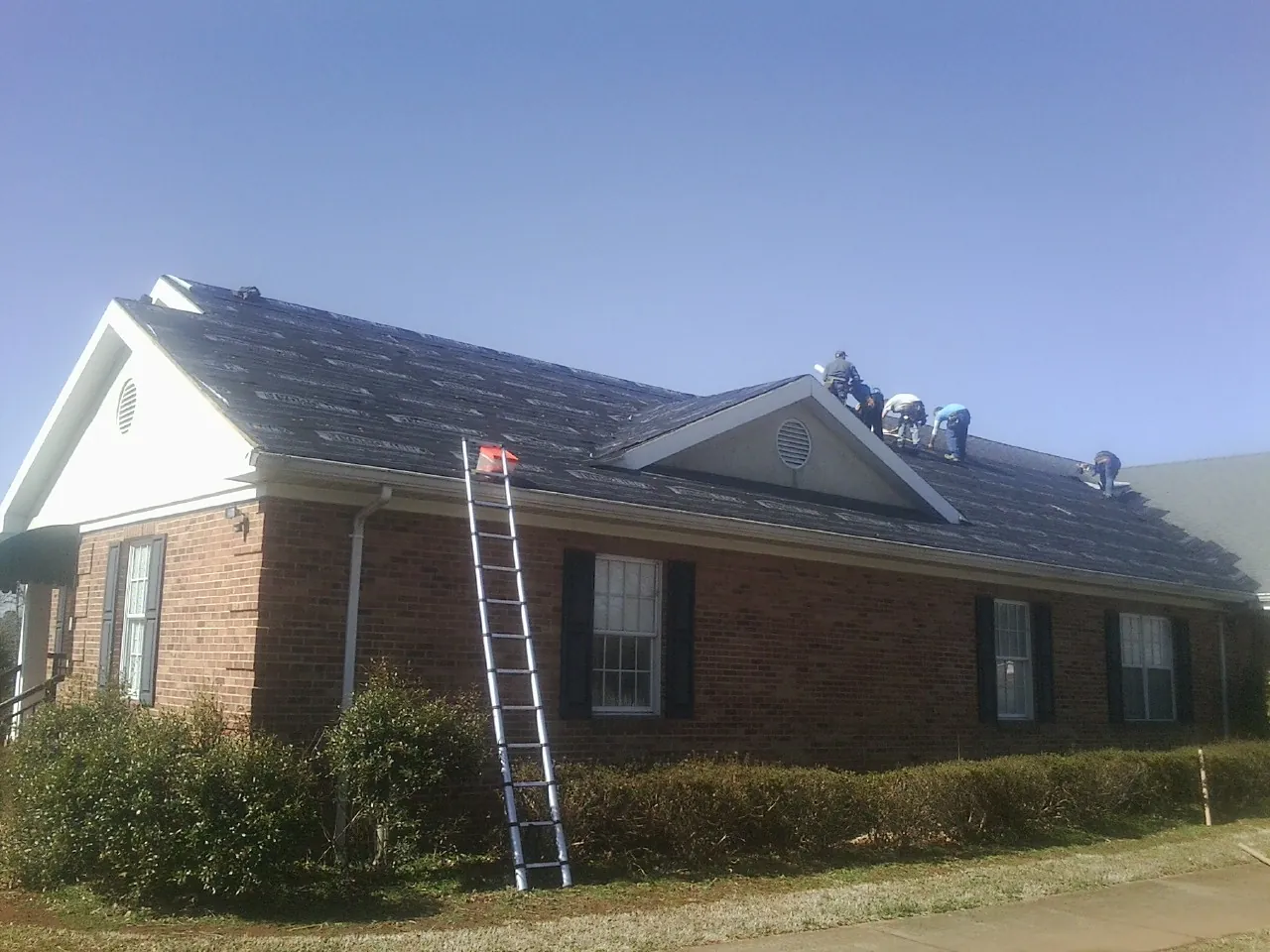 Crew tearing shingles off a brick ranch roof with a ladder set — Metal Roof Repair work by our crew in Wampsville