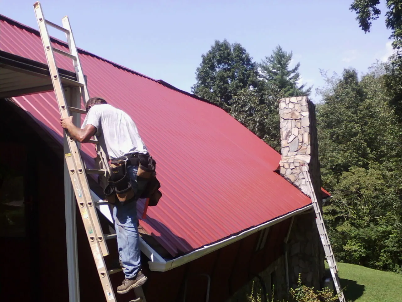 Roofer on a ladder beside a red metal roof and stone chimney — Soffit & Fascia Repair by Roofing Brothers in Wampsville
